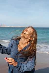 Happy female in wet shirt standing on sandy beach near sea while holding photo camera on sunny day