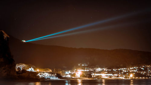 Laser light by illuminated buildings and sea against sky at night