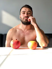 Shirtless man sitting with pomegranate and orange on table at home