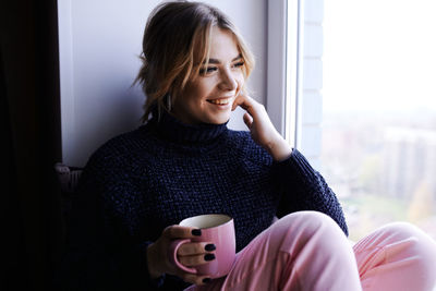 Smiling woman holding coffee cup sitting by window at home