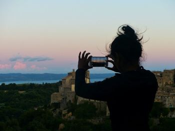 Silhouette woman photographing with smart phone against sky during sunset