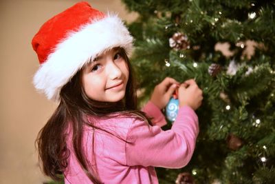 Portrait of smiling girl wearing santa hat while decorating christmas tree at home