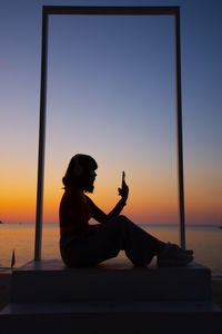 Woman sitting on mobile phone against sky during sunset