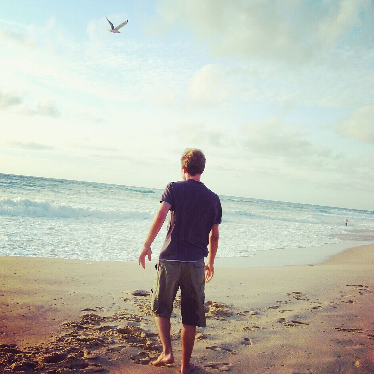 REAR VIEW OF MAN STANDING ON BEACH AGAINST SKY
