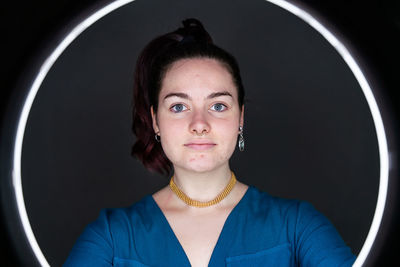 Charming female illuminating face with circle lamp while standing in dark studio and looking at camera