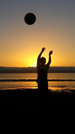 Silhouette boy playing with ball at beach against sky during sunset