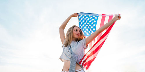 Young millennial blonde woman standing with american flag next to blue sky. flag of the united state