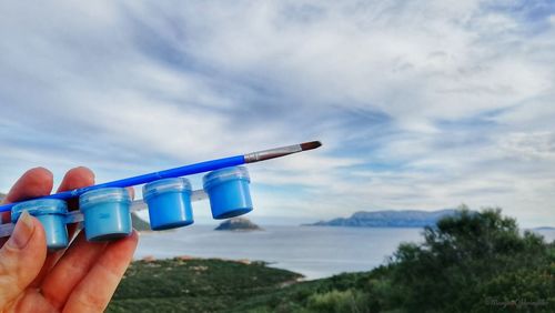 Close-up of person holding paint and paintbrush against sky