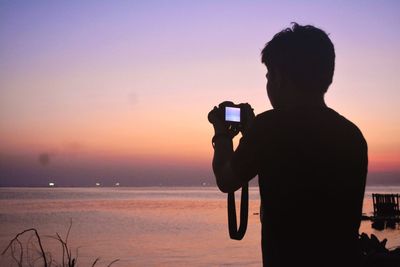 Silhouette man photographing against sea during sunset