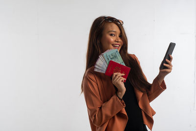 Portrait of young woman holding smart phone while standing against wall