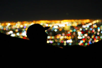 Defocused image of silhouette man standing in illuminated city against sky at night