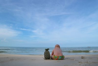 Rear view of baby boy sitting by pineapple at shore of beach