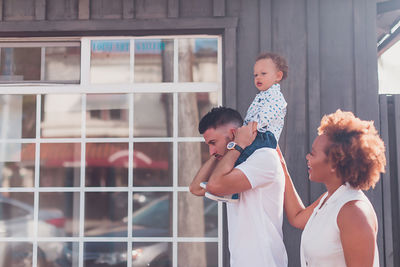 Dad carrying baby on shoulders with mom smiling in front of big window