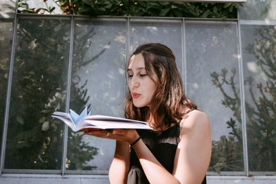 Young woman blowing origami crane on book by greenhouse