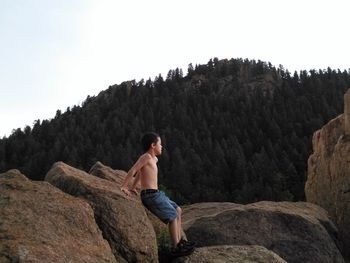 Full length of shirtless boy leaning on rock against mountain