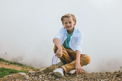 Portrait of smiling boy sitting on rock against sky