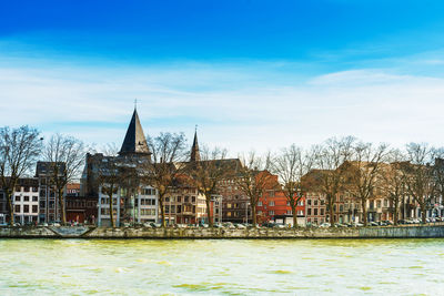 View of buildings by river against cloudy sky during sunny day