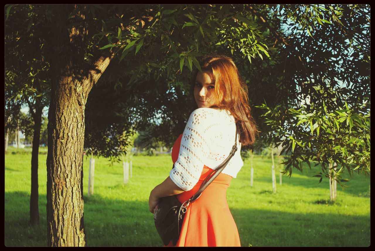 PORTRAIT OF SMILING YOUNG WOMAN STANDING BY TREE