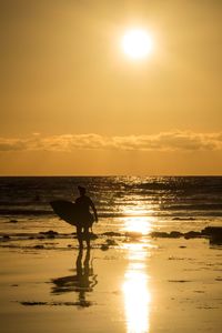 Silhouette person with surfboard walking on shore at beach against sky during sunset