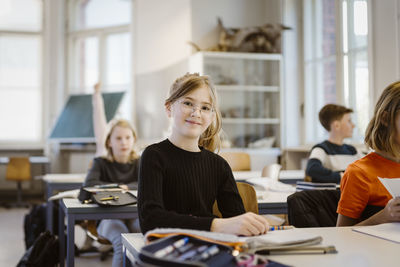 Portrait of smiling schoolgirl sitting at desk with friend in classroom