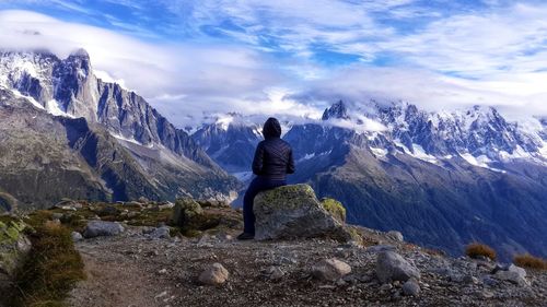Rear view of woman sitting on rock while looking at mountains against sky