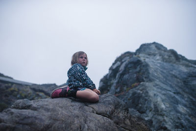 Low angle view of carefree girl sitting on rock against mountains and sky