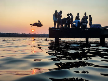 Man jumping in lake with friends standing on pier during sunset