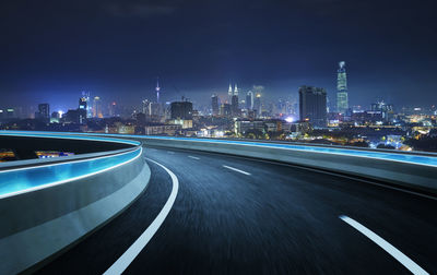 Illuminated road by buildings against sky at night