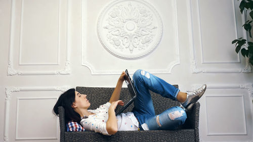 Young woman, girl, brunette, in white shirt and jeans, lying on a sofa, small couch, with laptop. 