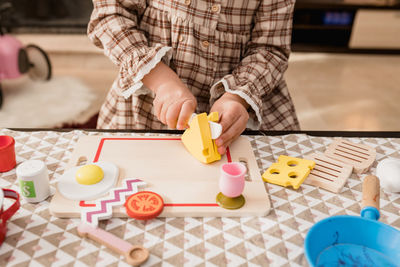 Crop unrecognizable kid in checkered dress with toy knife cutting cheese on chopping board while playing in house