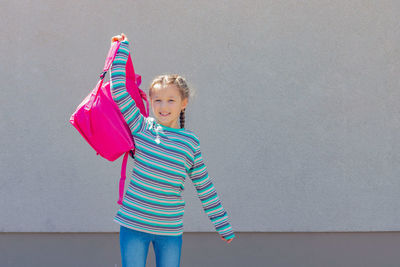 A cute little girl smiles and holds a pink backpack on her outstretched hand.