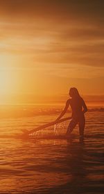 Silhouette young woman surfing on sea against cloudy sky during sunset