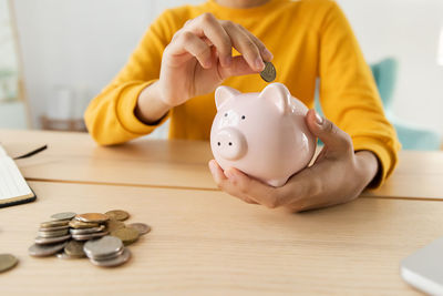 Midsection of woman putting coin in piggy bank on table