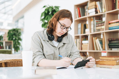 Young teenager girl college student in glasses doing homework with mobile phone at modern library 