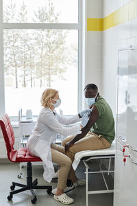 Doctor giving vaccine to patient sitting on bed in clinic