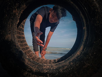 Young man gesturing while looking through tire against sky