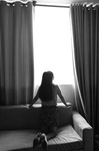 Rear view of young woman kneeling on sofa by window