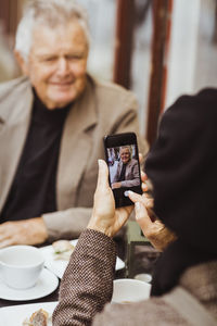 Woman photographing senior man through smart phone at sidewalk cafe