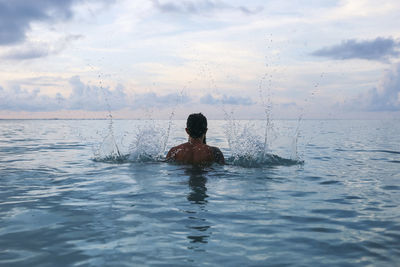 Rear view of shirtless man splashing water in sea against cloudy sky
