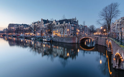 Illuminated bridge over river in city against sky at dusk