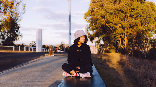 Young woman sitting on road amidst trees against sky