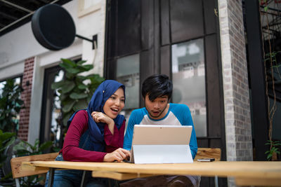 Low angle view of happy couple using digital tablet while sitting at sidewalk cafe