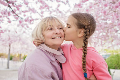 Girl kisses her grandmother near blooming branch of sakura during walking in spring garden