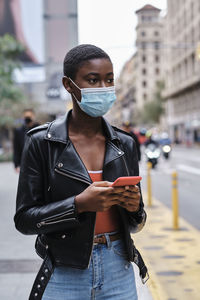 Young woman wearing protective face mask using mobile phone while standing in city