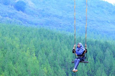 Portrait of young woman swinging against trees