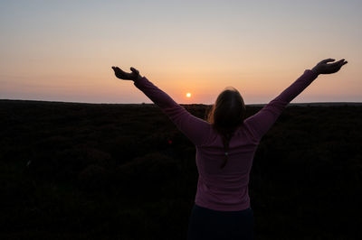 Rear view of woman with arms raised standing against sky during sunset