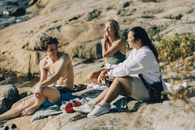 Happy multiracial male and female friends spending leisure time sitting on rock at sunny day