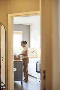 Side view full length of mature woman unpacking package while standing in kitchen seen through doorway at home