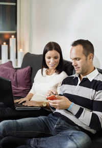 Couple using mobile phone and laptop on sofa at home