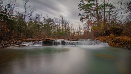 Panoramic long-exposure scenic view of a small waterfall in a forest setting with a moody sky